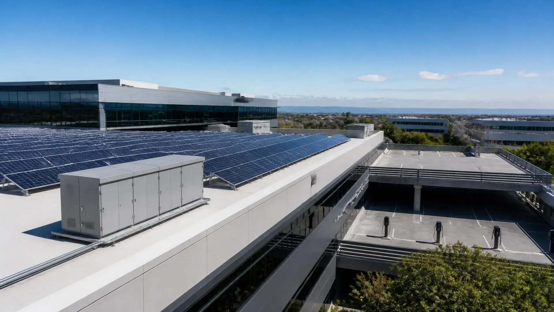 Elevated daylight view of a commercial business park with rooftop solar arrays, a rooftop battery enclosure, and charging pedestals on the parking deck below.
